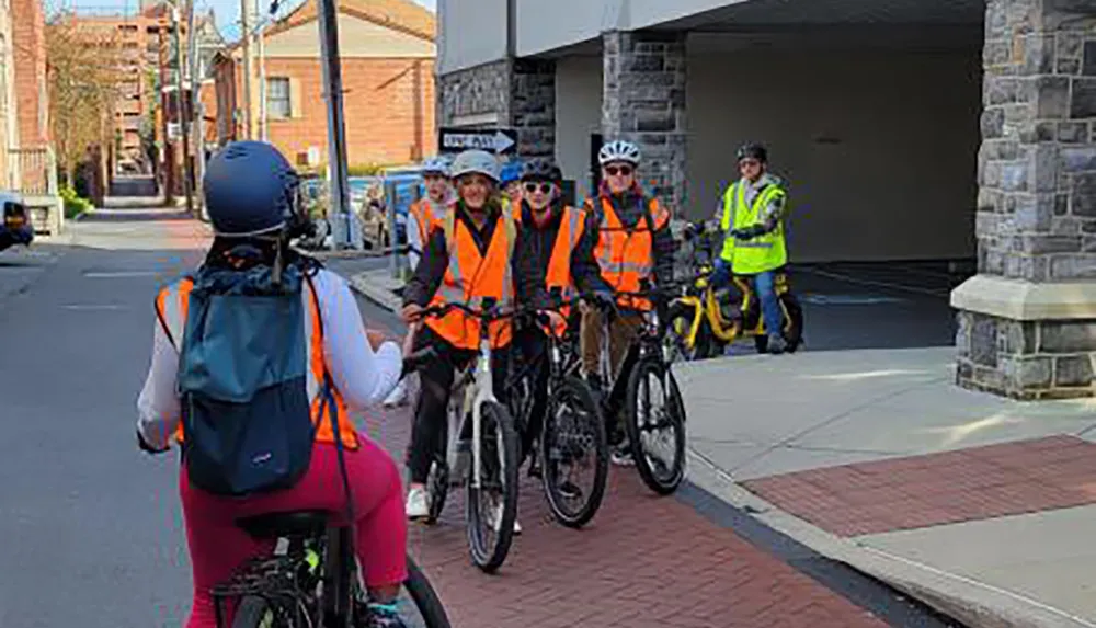 A group of cyclists wearing safety vests and helmets are gathered on a city street possibly preparing for a group ride or participating in a cycling event