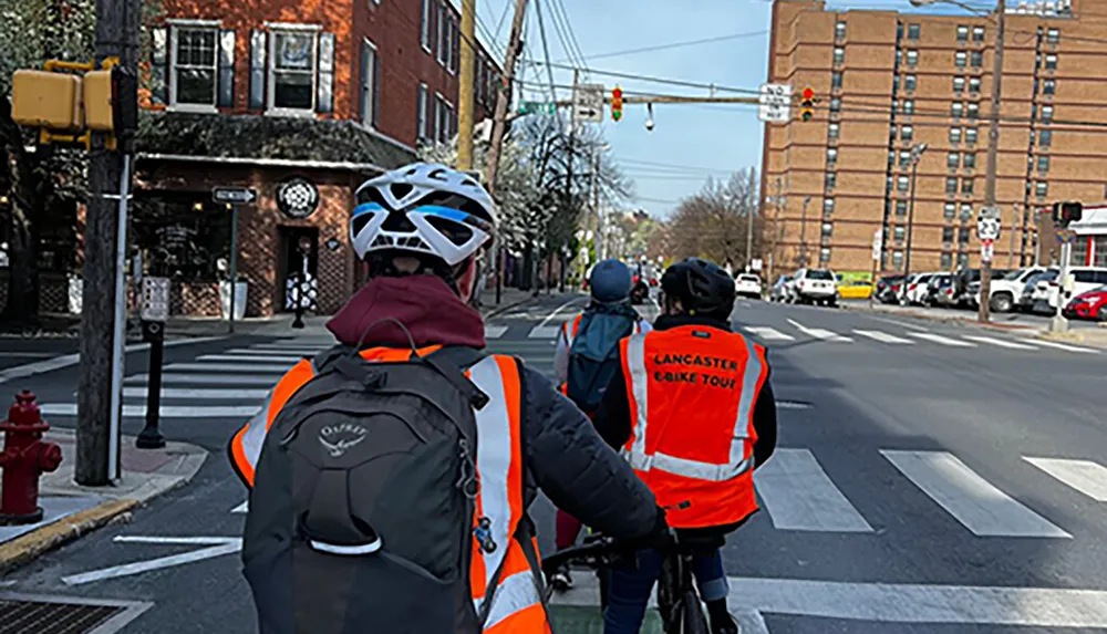 A group of cyclists wearing safety gear and high-visibility vests is waiting at a traffic intersection in an urban area