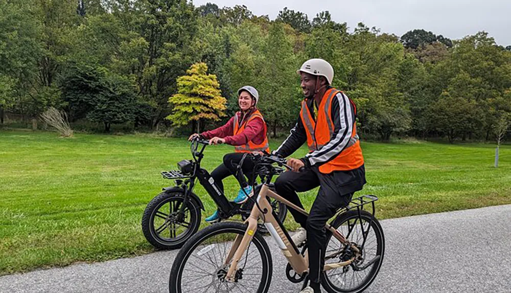 Two people wearing safety vests and helmets are riding bicycles on a paved path with greenery around them