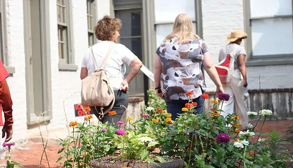 A group of people is walking through a sunny alleyway flanked by colorful flowers and white buildings