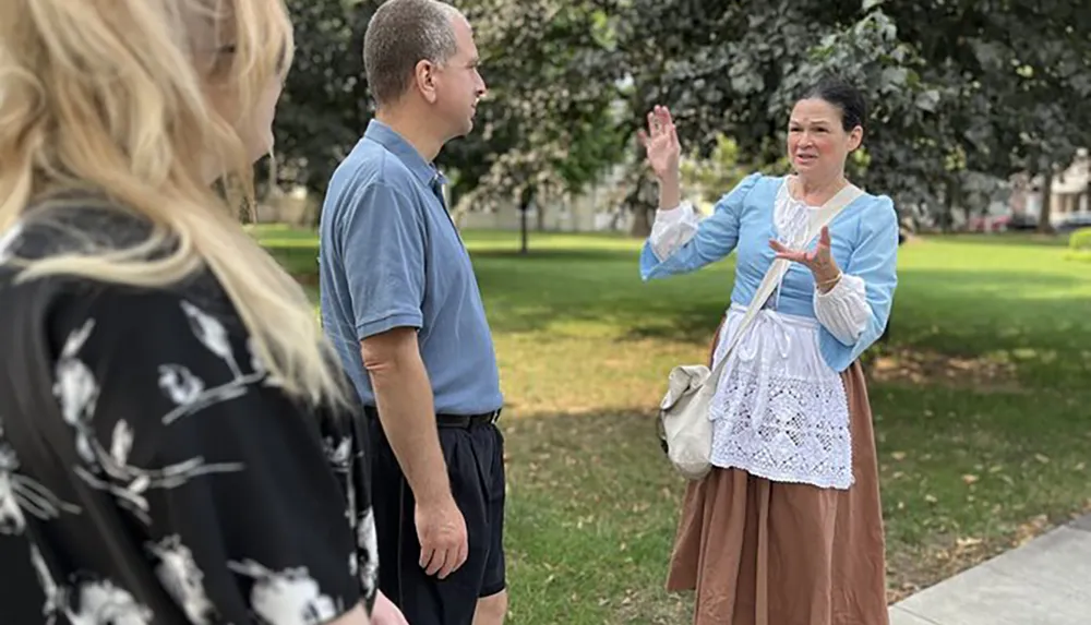 A woman dressed in historical costume is engaging in conversation with a man and a woman in a park setting