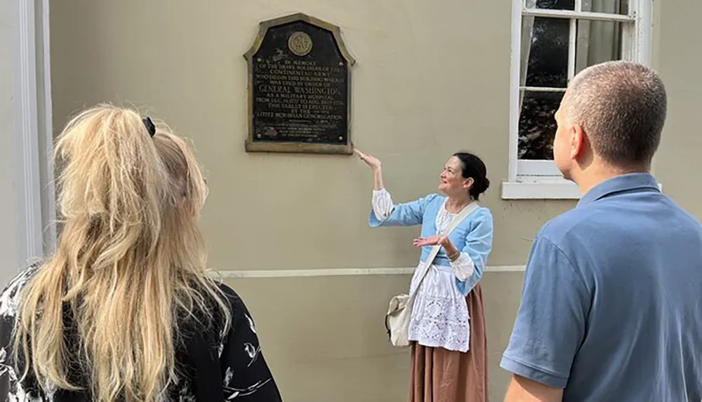 A person dressed in historical costume appears to be giving a tour or presentation to a small group in front of a commemorative plaque on a buildings exterior wall