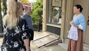 A woman in historical costume is speaking to two attentive listeners on a sidewalk, possibly conducting a historical tour.