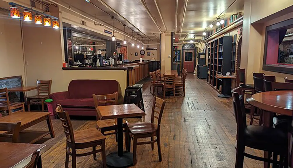 The image shows an empty warmly lit pub interior with wooden tables and chairs a bar counter to the right and shelves lined with books against the far wall