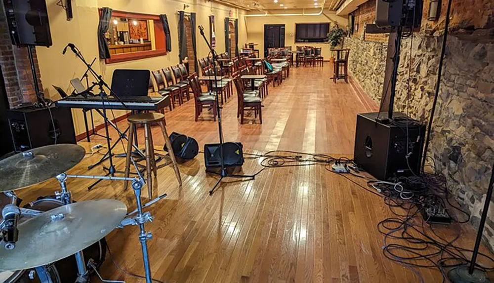 The image shows an empty venue set up for a musical performance featuring chairs arranged for an audience a stage area with microphones a keyboard drum set speakers and various cables