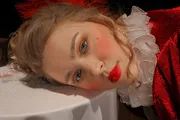 A person with glamorous makeup and a red costume with a frilly white collar is lying down, gazing toward the camera.