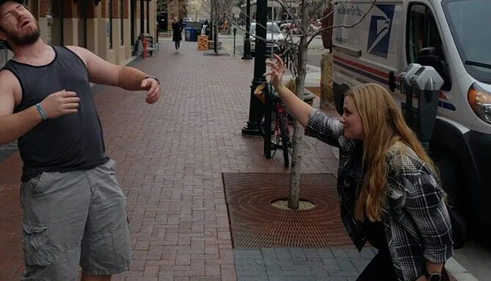 A man appears to be dramatically recoiling from a playful punch thrown by a smiling woman on a city sidewalk