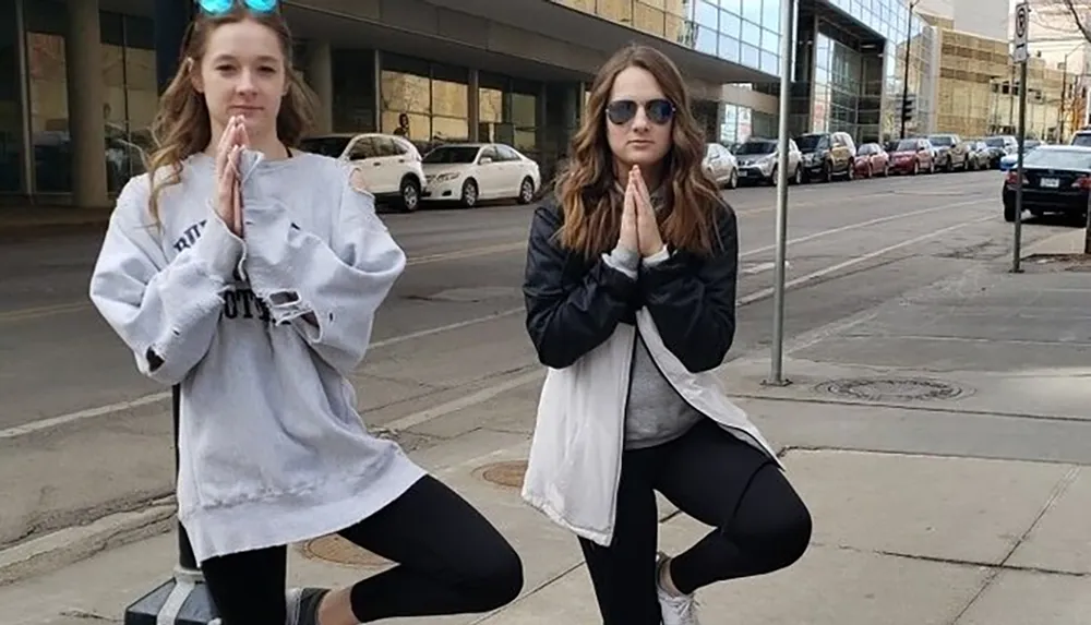 Two individuals are playfully posing with a yoga gesture on a city sidewalk