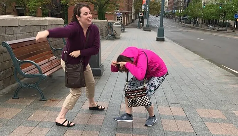 A woman in a pink jacket is doubled over in laughter on a city sidewalk while another woman in a purple cardigan appears to be playfully throwing something at her