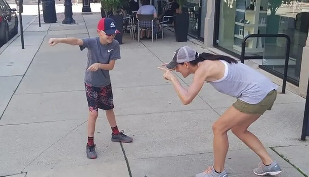 A child and an adult are engaging in a playful dab pose on a sunny sidewalk