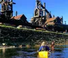 Two individuals are kayaking on a sunny day with a backdrop of lush greenery and an industrial structure