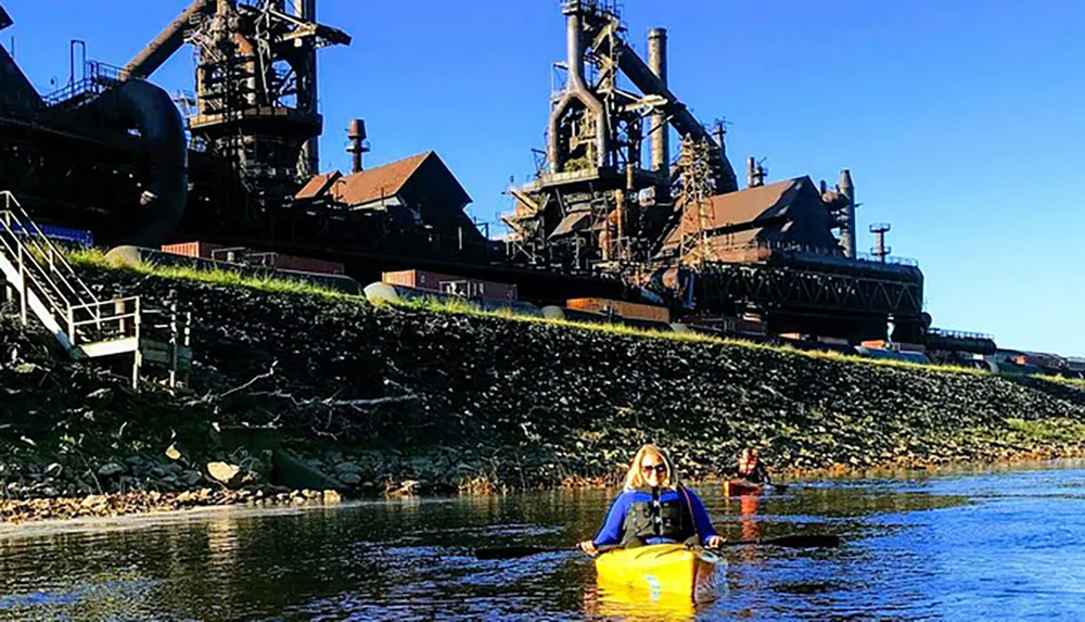 A person is kayaking on a river with a large industrial structure which appears to be a steel mill in the background under a clear blue sky