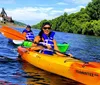Two individuals are kayaking on a sunny day with a backdrop of lush greenery and an industrial structure