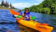 Two individuals are kayaking on a sunny day with a backdrop of lush greenery and an industrial structure.