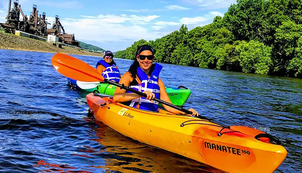 Two individuals are kayaking on a sunny day with a backdrop of lush greenery and an industrial structure