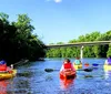 Two individuals are kayaking on a sunny day with a backdrop of lush greenery and an industrial structure