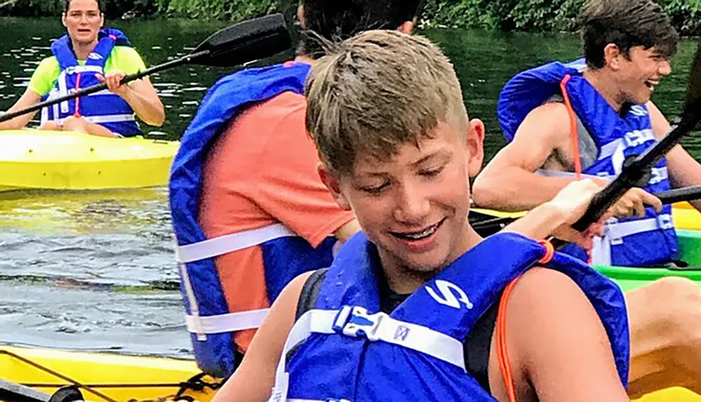 A group of people are enjoying kayaking on a body of water with two individuals in focus smiling and paddling their kayaks