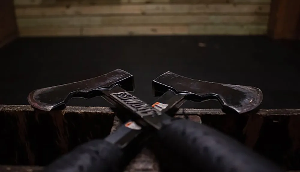 Two axes with black handles are placed in an X formation on a wooden surface against a blurred background