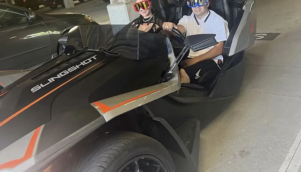 Two people are seated in an open three-wheeled vehicle labeled Slingshot wearing casual clothing and protective eyewear seemingly ready for a ride