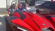 Two people are sitting in a red Polaris Slingshot vehicle parked in a residential driveway on a sunny day.