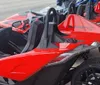 The image shows a red and black three-wheeled vehicle resembling a mix between a car and a motorcycle parked on an asphalt surface with buildings in the background