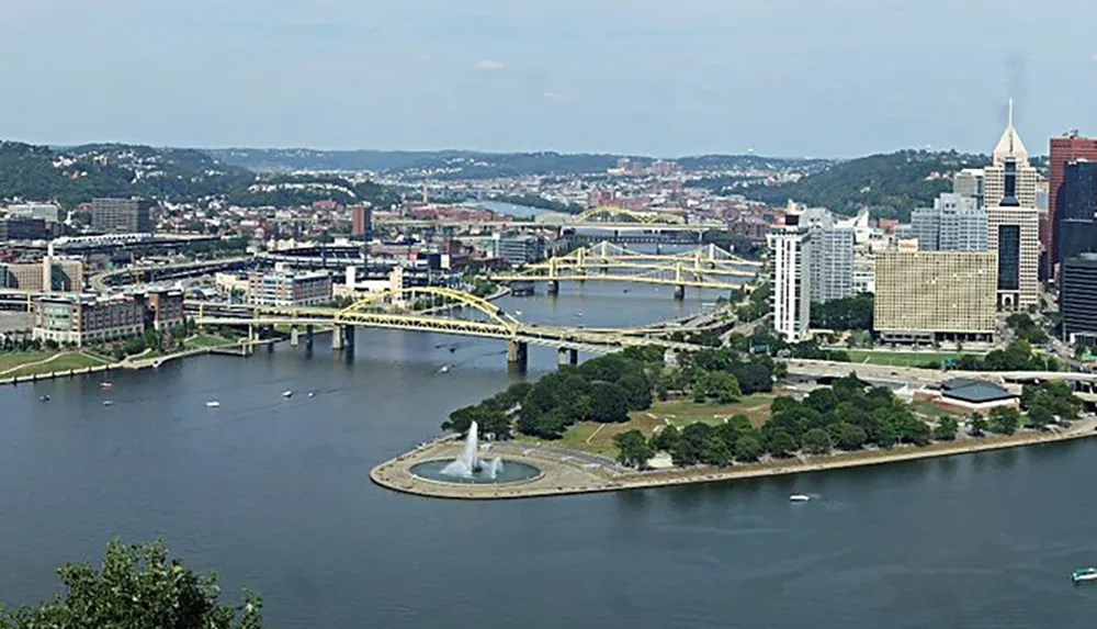 The image shows a panoramic view of a city with multiple bridges spanning a river a fountain in a park at the forefront and a mix of green spaces and urban architecture