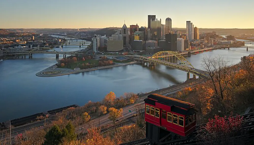 The image captures a hillside view of Pittsburghs skyline with its iconic yellow bridges over the Allegheny River and the Duquesne Incline in the foreground at dawn or dusk
