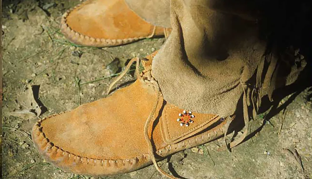 The image shows a close-up of a persons feet wearing traditional suede moccasins with fringe and beadwork detailing on a natural ground surface