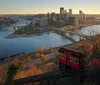 A red funicular car descends along a track overlooking a panoramic view of a city with skyscrapers bridges and rivers during sunset