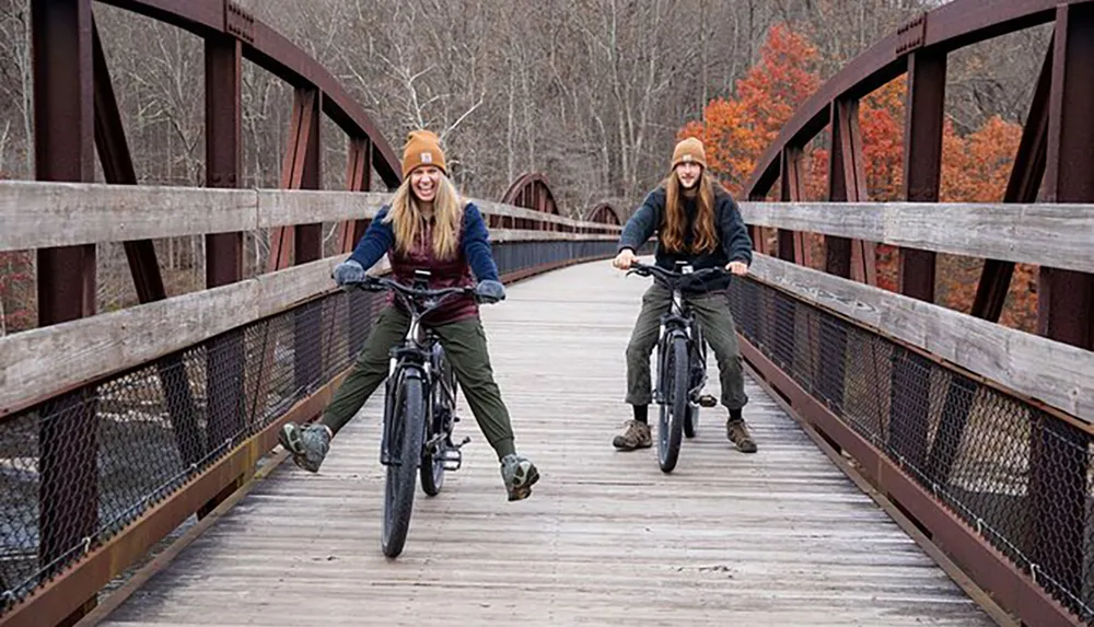 Two people are joyfully riding bicycles on a wooden bridge surrounded by autumn foliage