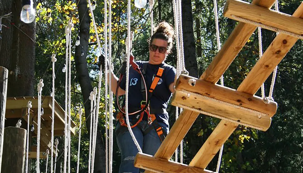 A person is navigating a rope and wooden plank obstacle course among the trees wearing safety harness equipment