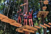 Three individuals are harnessed and smiling on an outdoor high ropes adventure course among the trees.