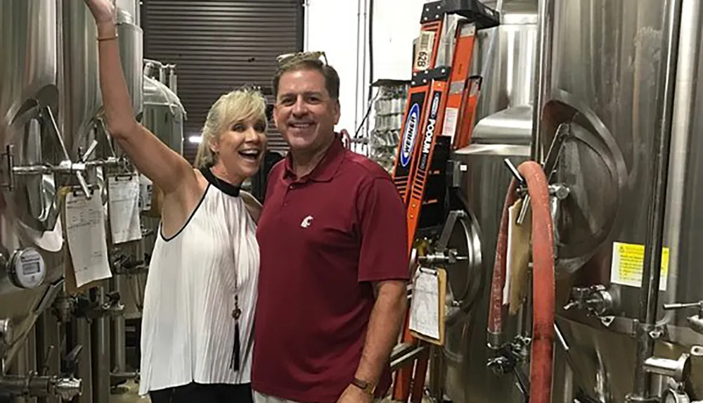 A smiling man and woman pose in front of stainless steel brewing equipment suggesting a setting in a brewery