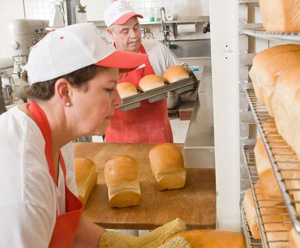 Two bakers are working in a bakery with one arranging freshly baked loaves of bread on a wooden board while the other looks on with a tray of buns
