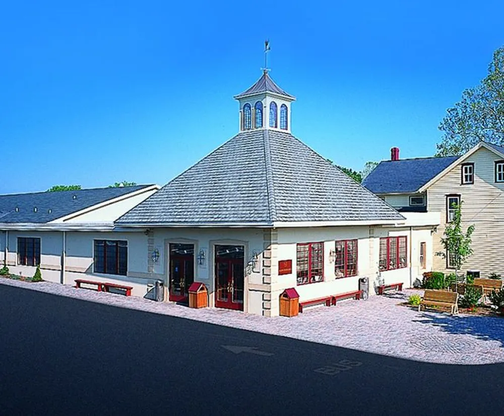 The image shows a quaint building with a distinct octagonal shape and a cupola featuring a deep red door and surrounded by a paved area with benches