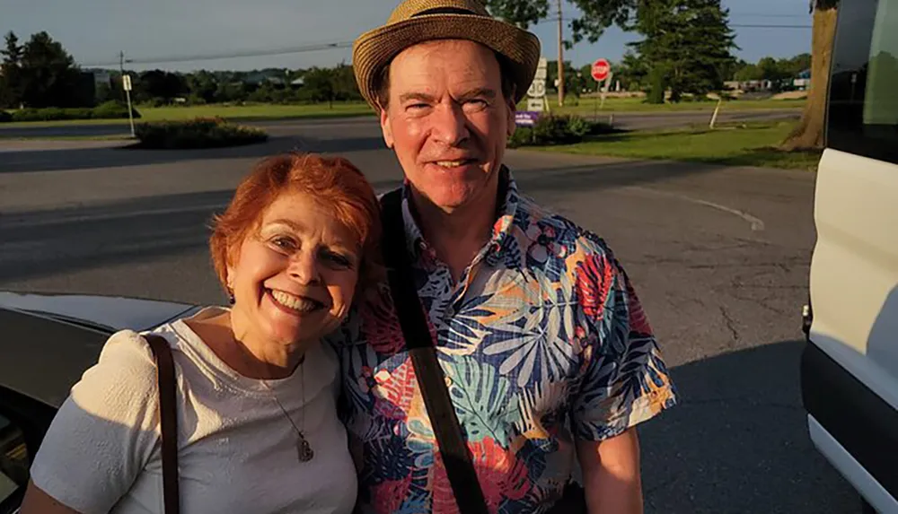 A smiling couple is posing for a photo in casual summer attire with the woman leaning towards the camera and the man wearing a straw hat and tropical shirt