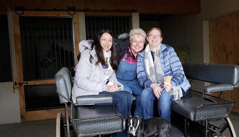 Three women and a dog are smiling for the camera inside a stable with a horse visible in the background