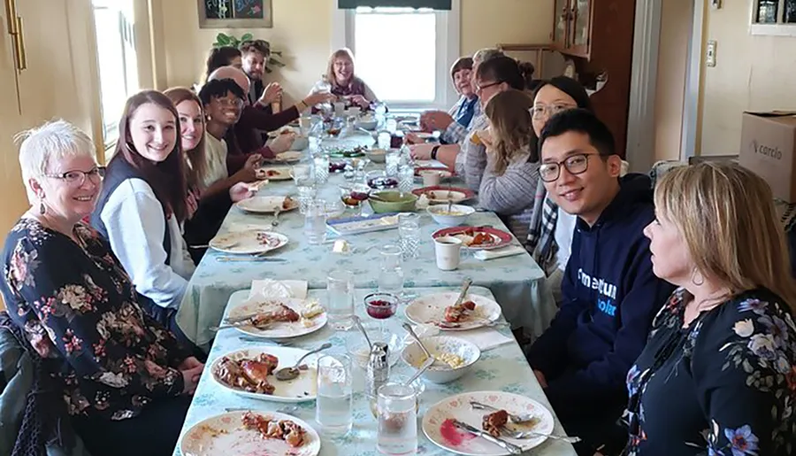 A group of people is smiling and engaging with each other around a table with the remains of a meal, indicating a social gathering such as a family dinner or a festive occasion.