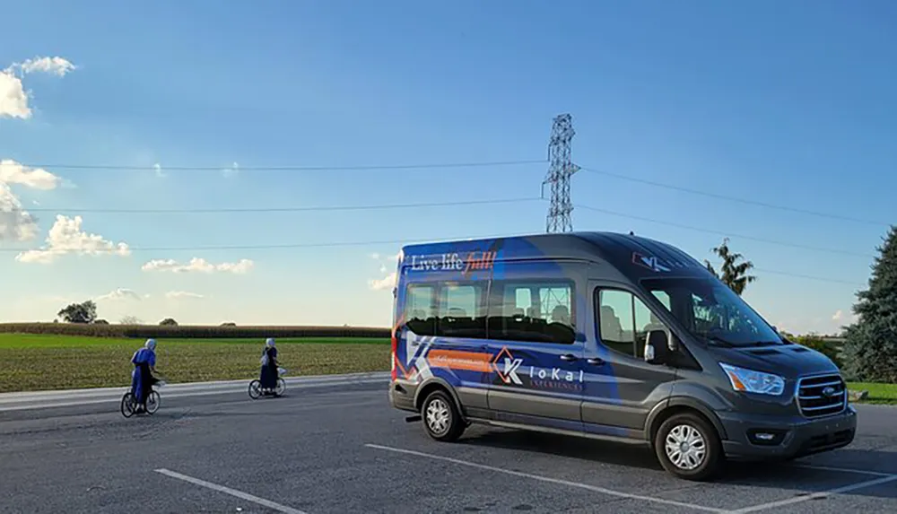 A shuttle bus branded Live life Fully is parked on the side of the road as two individuals on bicycles ride past in a rural setting