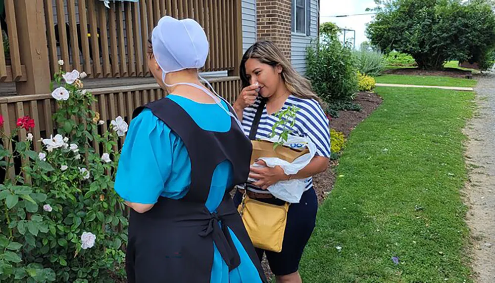 Two women are conversing outdoors one dressed in traditional attire with a head covering and the other in modern casual wear with a garden setting in the background