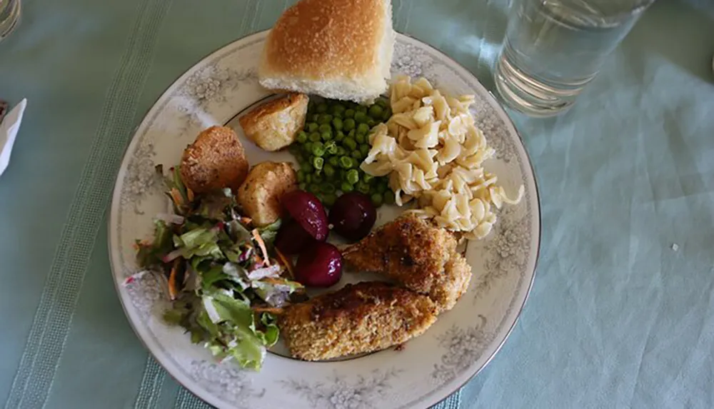 The image shows a plate with a variety of foods including breaded chicken peas pasta mixed salad roasted potatoes beets and a slice of bread accompanied by a glass of water