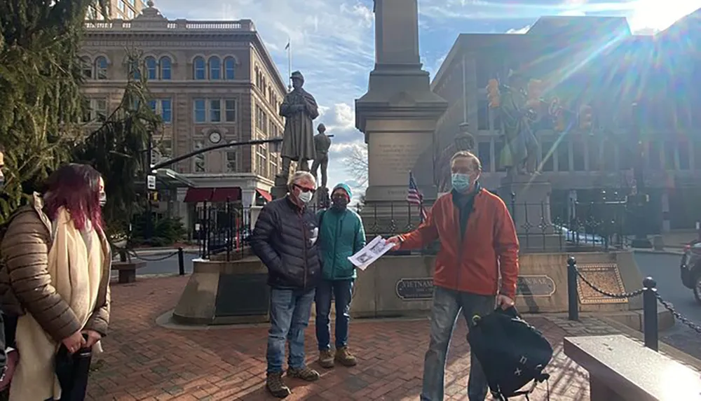 A group of individuals with masks stands near a statue in a town square with one person handing a paper to another under a bright sun