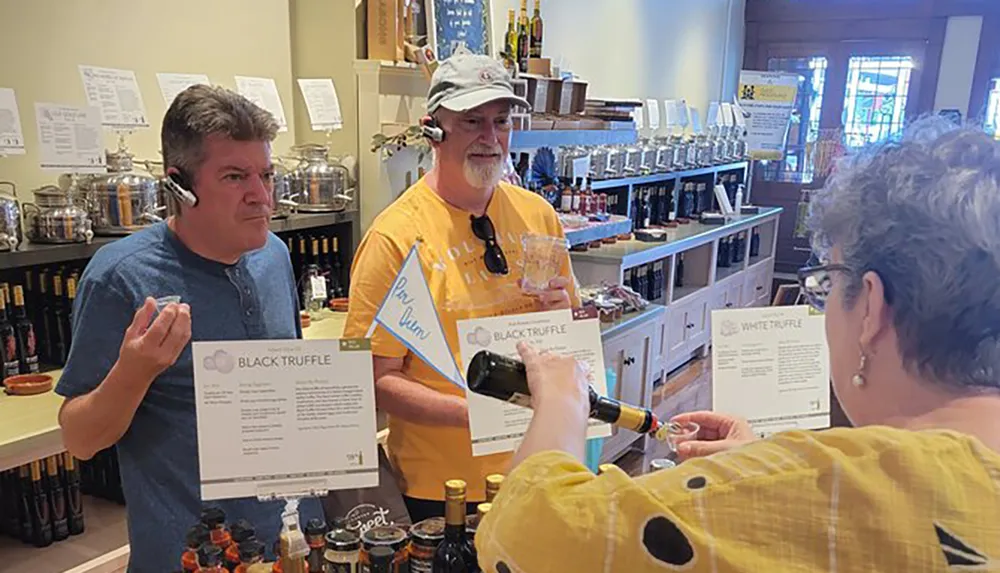 In the image two individuals appear to be participating in an oil and vinegar tasting at a specialty store with one man evaluating a product and another holding a tasting glass and bottle with informational signs about black and white truffle flavors displayed in front of them