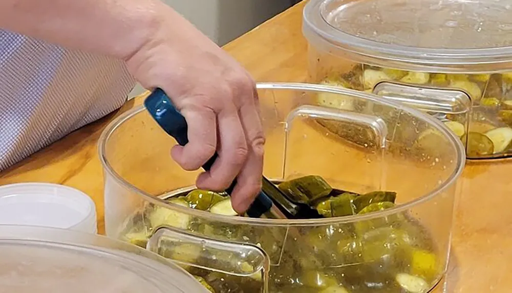 A person is using tongs to pick up some pickles from a clear plastic container