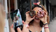 A person is playfully holding up two colorful doughnuts to their eyes like glasses while another person takes their photo with a smartphone.