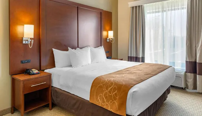 The image shows a neatly arranged hotel room with a large bed a bedside table wall-mounted lamps and a window with curtains