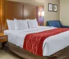 The image shows a neatly made hotel room with a large bed red and white bedding a side table wall-mounted lights and an armchair creating a cozy and welcoming atmosphere