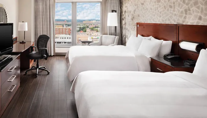 This image shows a neatly arranged hotel room with two beds a desk with an office chair and a view of the city from the window