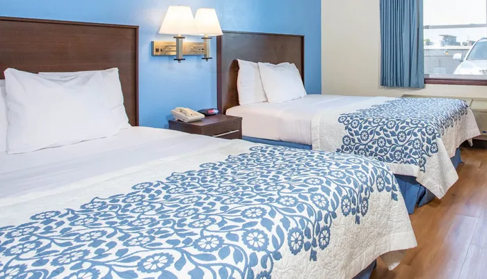 The image shows a neatly arranged hotel room with two twin beds covered with blue and white patterned bedspreads a nightstand with a phone and a window that allows natural light into the room