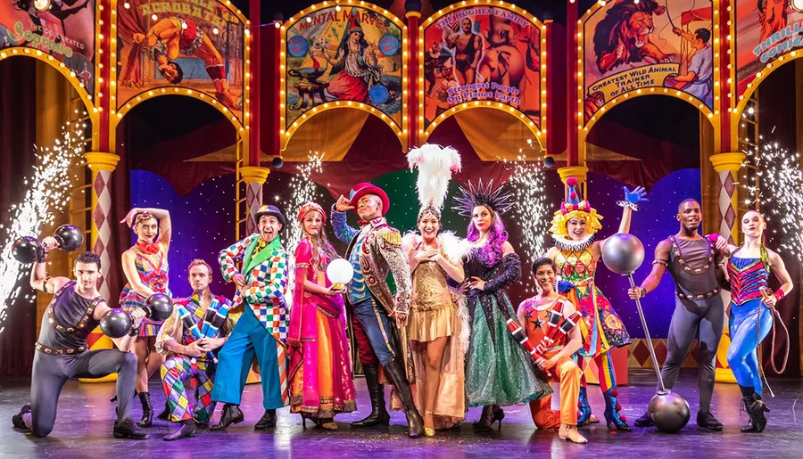 A group of performers in vibrant costumes poses on stage amongst sparklers, showcasing a variety of circus acts against a colorful backdrop of vintage circus posters.